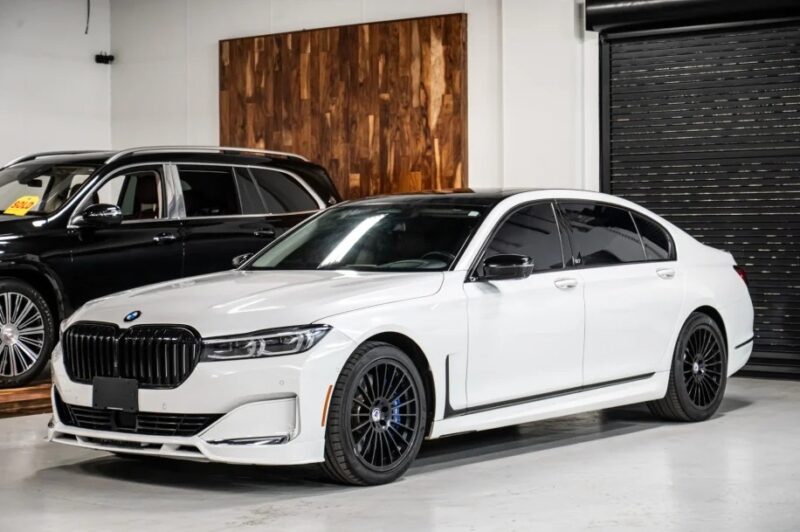 A white luxury sedan is parked indoors, exuding elegance beside a sleek black vehicle, with a wooden panel adorning the wall in the background.