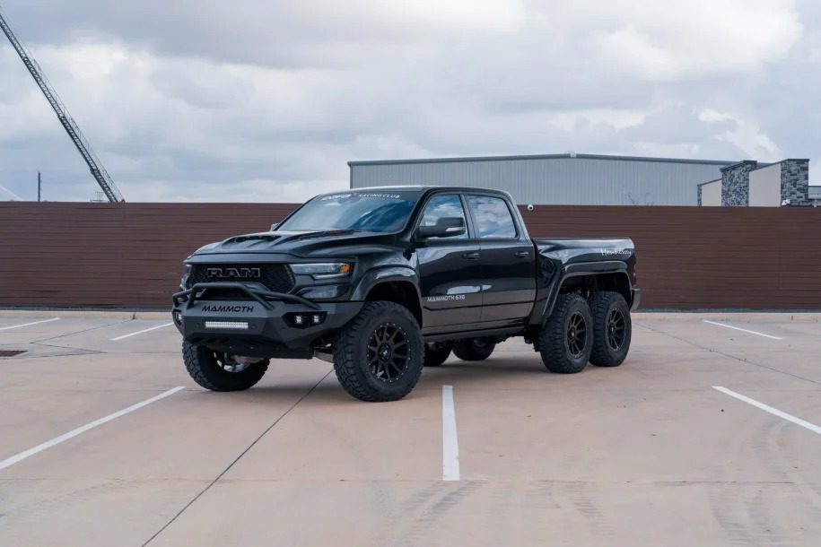 A striking black 6x6 truck, sure to turn heads, is parked in a lot under a cloudy sky with industrial buildings looming in the background.