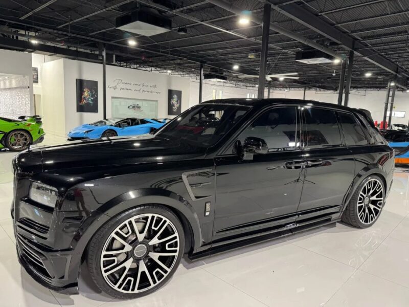 The pinnacle of automotive elegance, a black Rolls-Royce Cullinan with custom rims, is parked in a glossy showroom, surrounded by other high-end cars in dazzling colors.