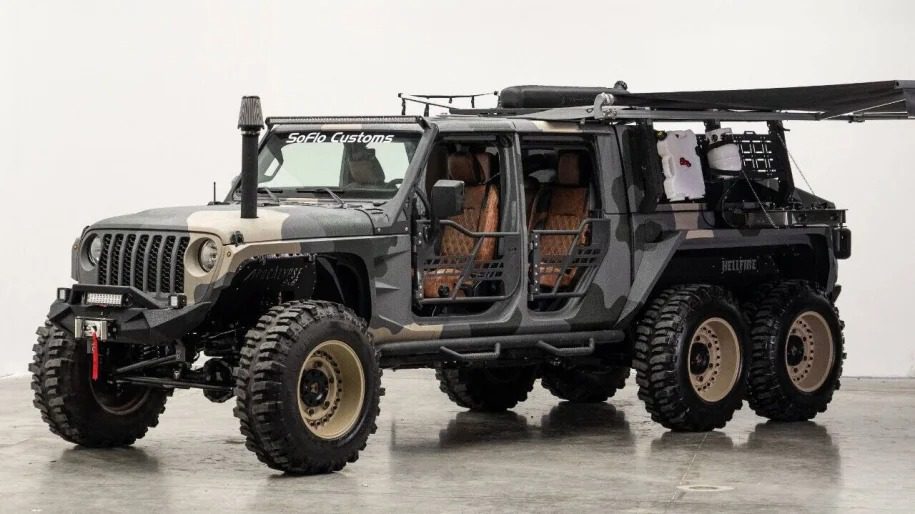 A heavily modified 6x6 Jeep with open doors, a winch, and a rooftop awning is parked indoors, ready to turn heads.