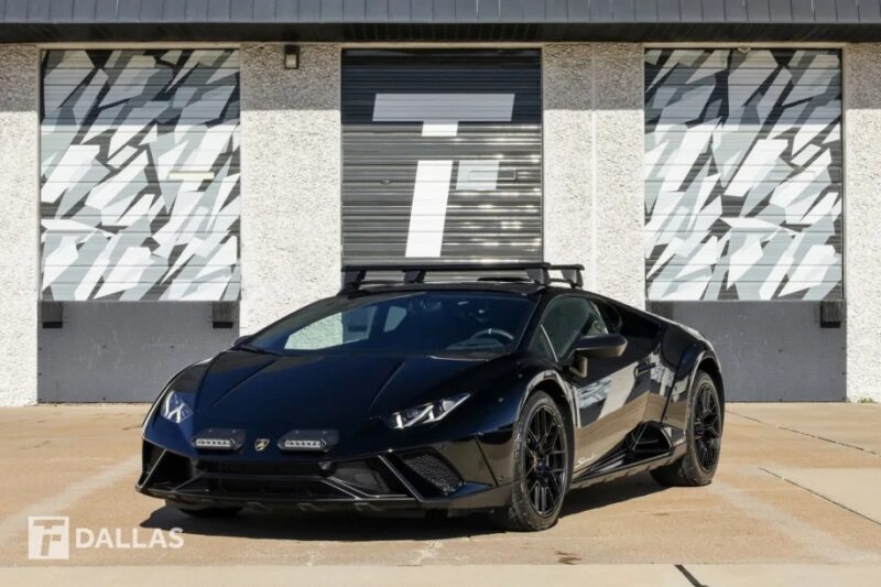 A sleek black sports car with roof racks is parked in front of a patterned garage door, proudly displaying the "T DALLAS" branding. If you're wondering how to buy Lamborghini models like this, or the adventurous Lamborghini Huracán Sterrato, this striking scene is sure to inspire.