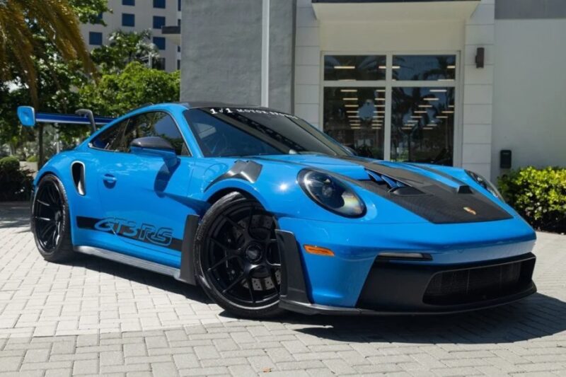 A 2023 Porsche 911 GT3 RS with black accents and a spoiler is parked on a gray paved area, near a building with large windows and green shrubs in the background.