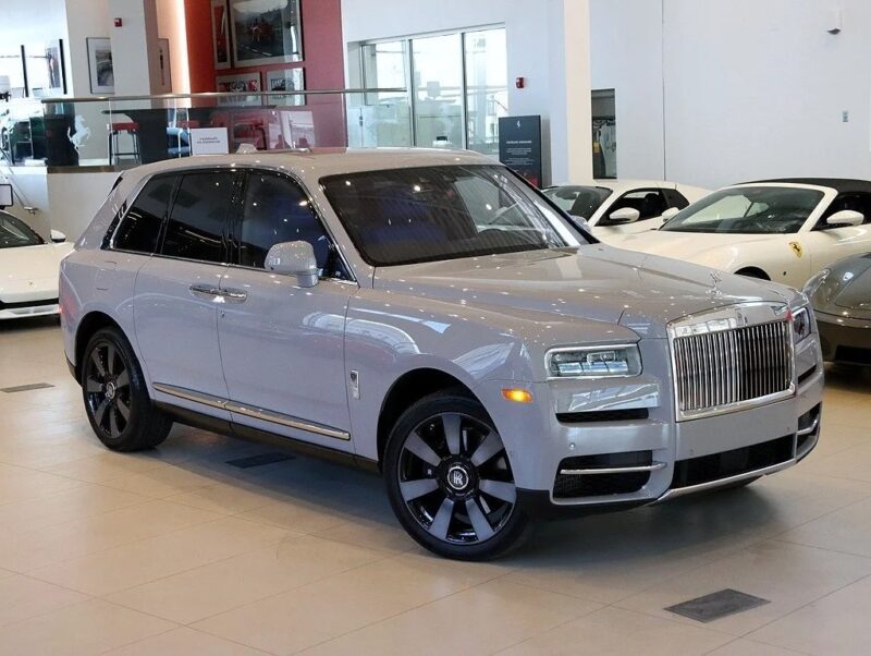 A silver luxury SUV, possibly the pinnacle of design, is parked indoors in a showroom, surrounded by other vehicles. The Rolls-Royce Cullinan boasts a prominent grille and dark alloy wheels, exuding an aura of elegance.