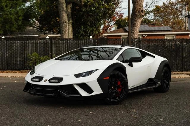 A sleek white Lamborghini Huracán Sterrato sports car is parked on the road, surrounded by trees and a building in the background.