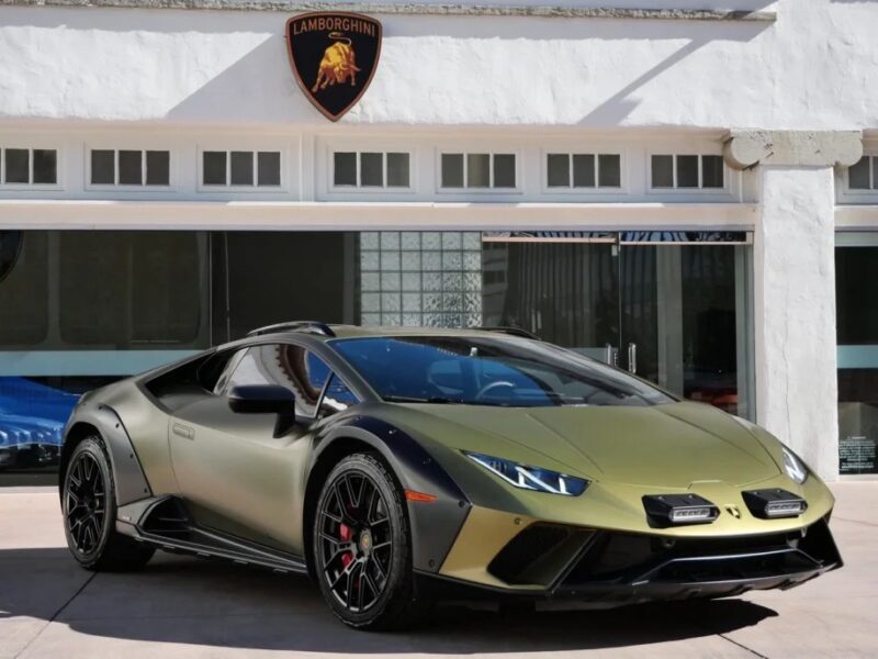 A green Lamborghini Huracán Sterrato with sleek black accents is parked in front of a garage, proudly displaying the iconic Lamborghini logo above. Considering a purchase? This adventurous Sterrato model offers an exhilarating experience that's hard to resist.