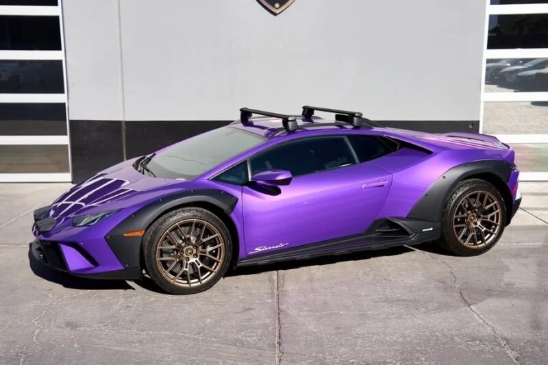 A Lamborghini Huracán Sterrato with striking purple paint, gold wheels, and roof racks is parked outside a building with garage doors. This eye-catching ride could be the perfect reason to explore a Lamborghini purchase and embrace the thrill of driving in style.