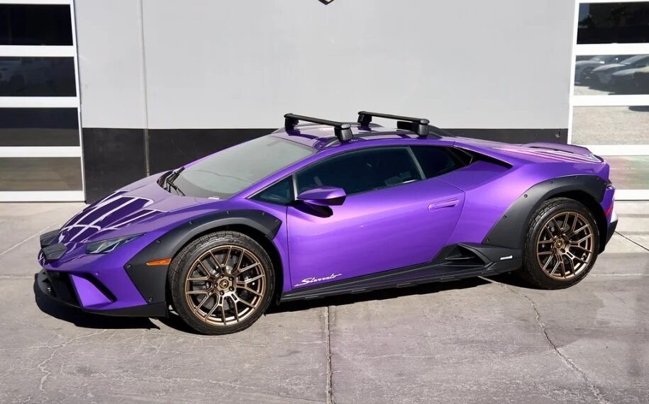 A Lamborghini Huracán Sterrato with striking purple paint, gold wheels, and roof racks is parked outside a building with garage doors. This eye-catching ride could be the perfect reason to explore a Lamborghini purchase and embrace the thrill of driving in style.