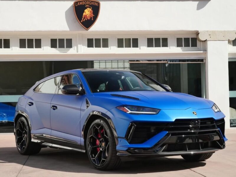 A sleek, blue Lamborghini Urus SUV is majestically parked in front of a building adorned with the iconic Lamborghini logo above its garage door.