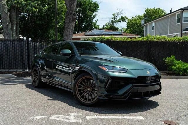 A dark green Lamborghini Urus, the renowned Italian Super SUV, is parked on a sunny street next to a black fence and trees. Houses are visible in the background.