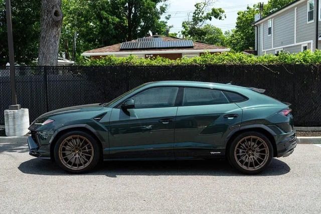 A dark green Lamborghini Urus with bronze wheels is parked on a street, with a fence and trees in the background, showcasing the elegance and power of this Italian Super SUV.