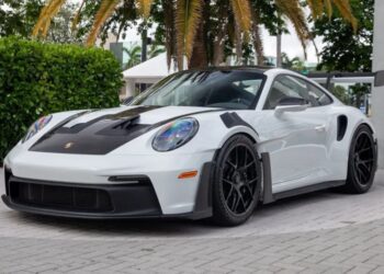 A 2023 Porsche 911 GT3 RS with black accents and a large rear spoiler is parked on a stone pavement, surrounded by greenery and palm trees.