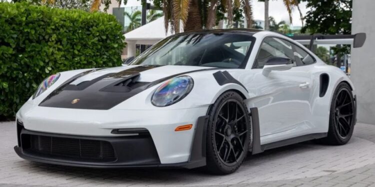 A 2023 Porsche 911 GT3 RS with black accents and a large rear spoiler is parked on a stone pavement, surrounded by greenery and palm trees.