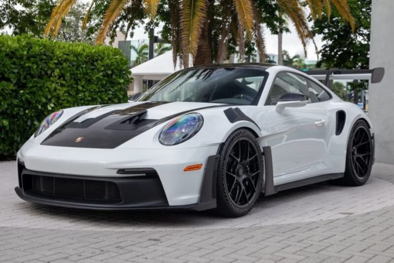 A 2023 Porsche 911 GT3 RS with black accents and a large rear spoiler is parked on a stone pavement, surrounded by greenery and palm trees.