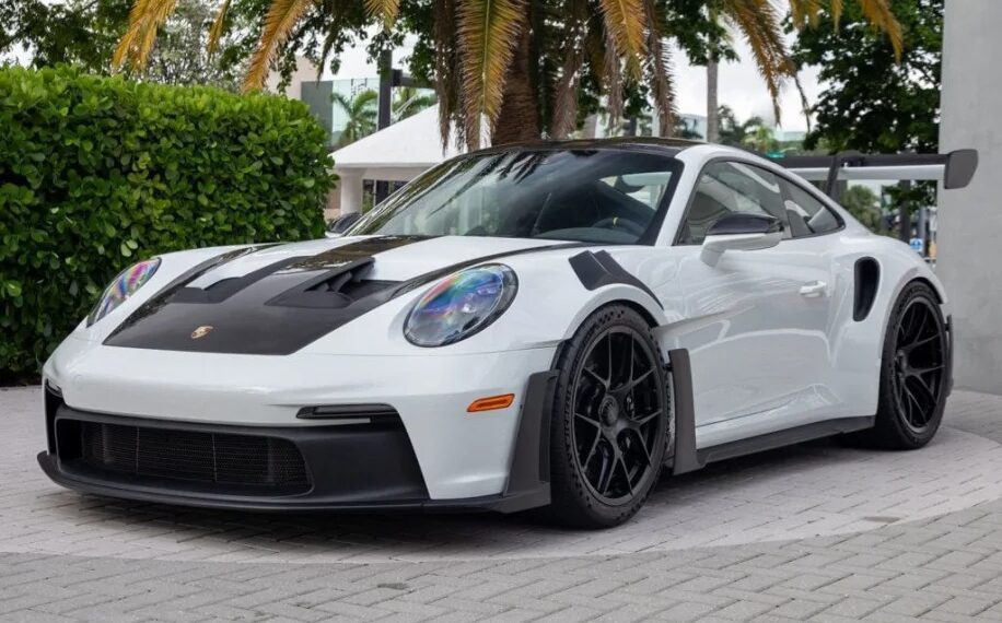 A 2023 Porsche 911 GT3 RS with black accents and a large rear spoiler is parked on a stone pavement, surrounded by greenery and palm trees.