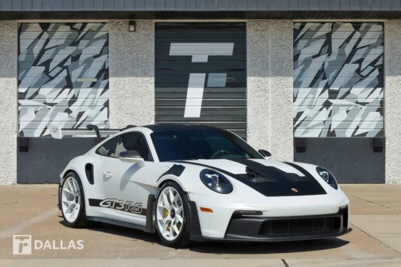 A sleek white 2023 Porsche 911 GT3 RS is parked in front of a building with patterned gray doors and the "T Dallas" logo.