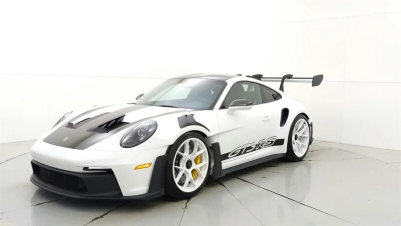 A sleek white Porsche 911 GT3 RS with black accents and a prominent rear wing is parked indoors, showcasing its 2023 design excellence.