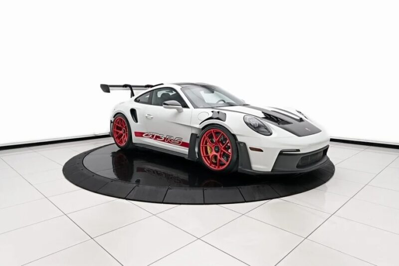 A stunning 2023 Porsche 911 GT3 RS, accented by red wheels and "GT3 RS" decals, is showcased on a circular platform in the showroom. Perfect for those eager to buy a blend of performance and style.