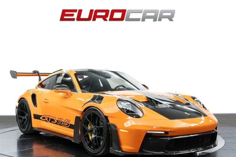 The 2023 Porsche 911 GT3 RS, in striking orange with sleek black accents and a large rear spoiler, flaunts its racing decals in the showroom. The "Eurocar" text above invites enthusiasts to explore and buy this high-performance marvel.