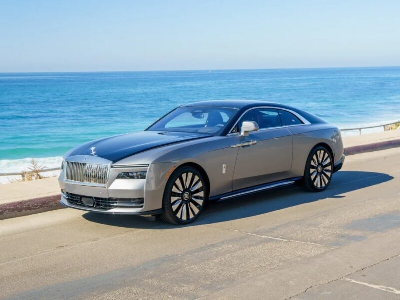 A silver, ultra-luxe Rolls-Royce Spectre, the epitome of an electric super coupe, is parked gracefully on a road by the ocean on a clear day.