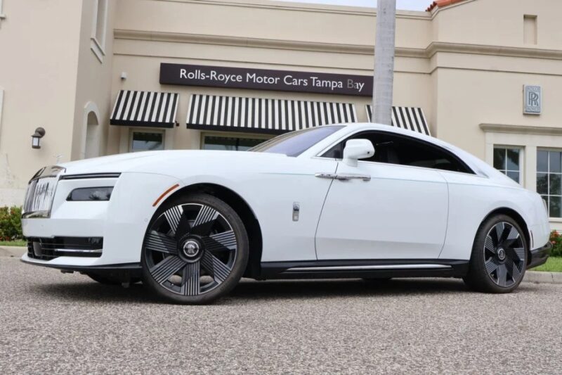 A white Rolls-Royce Spectre Super Coupe sits elegantly in front of a Rolls-Royce dealership with striped awnings, showcasing the epitome of electric ultra-luxe design.