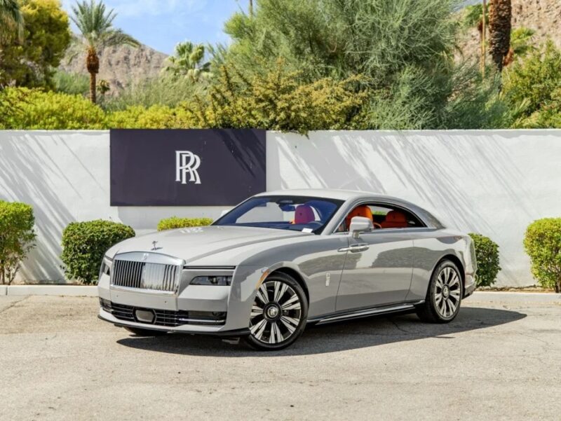 An ultra-luxe Rolls-Royce Spectre is gracefully parked in front of a white wall adorned with a black and white logo. The sleek, silver electric super coupe contrasts beautifully against the lush greenery and majestic mountains in the background.