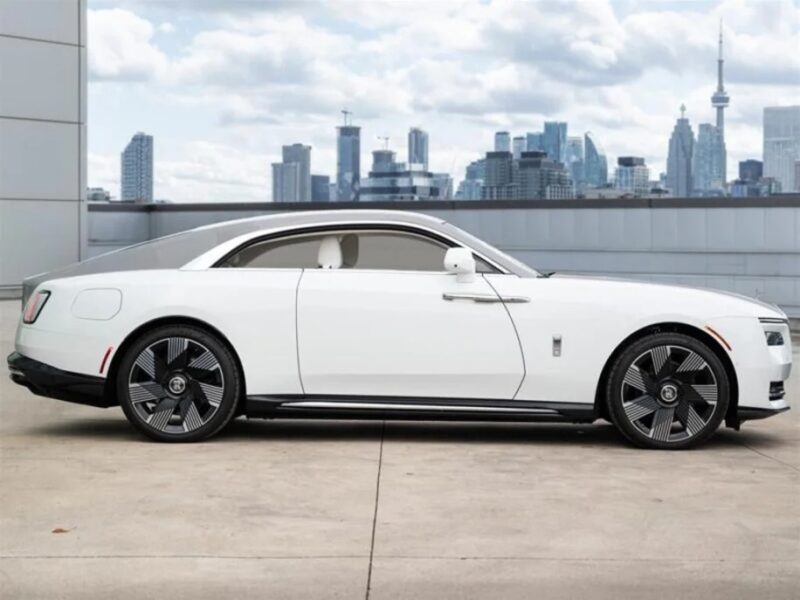 Side view of a sleek, white Rolls-Royce Spectre super coupe with black wheels parked on a rooftop, the electric ultra-luxe silhouette perfectly framed by the city skyline in the background.