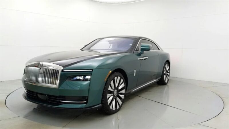 A green and black Rolls-Royce Spectre, an ultra-luxe electric super coupe, is displayed in a well-lit indoor studio with a pristine white background.