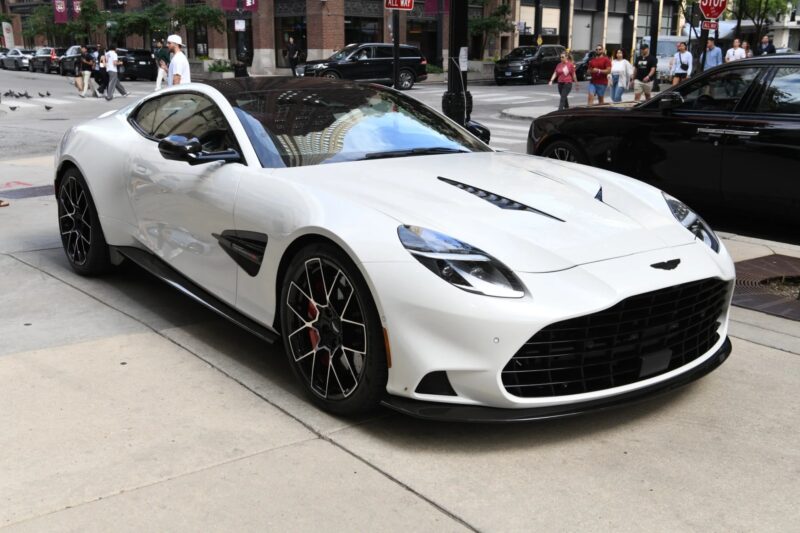 A white Aston Martin Vanquish V12 sports car is parked on a city street with pedestrians and buildings visible in the background.