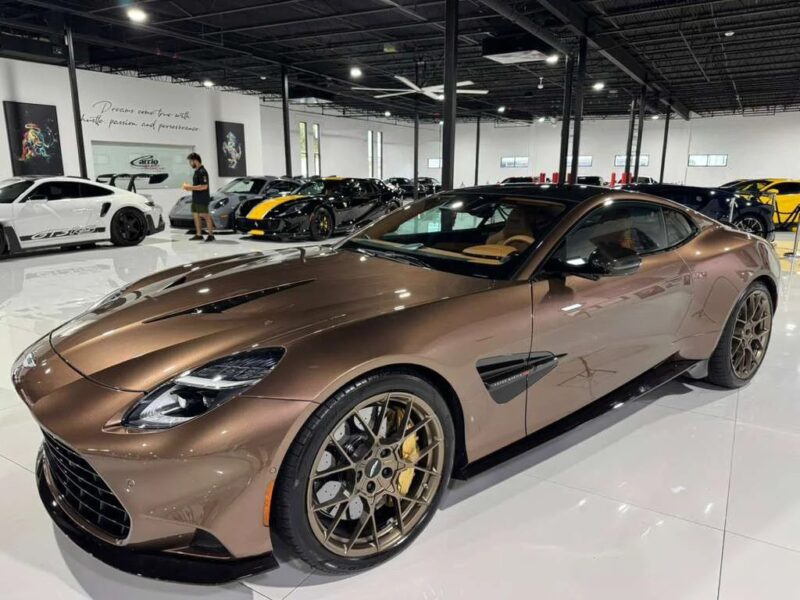 A metallic brown Aston Martin Vanquish V12 sports car is parked in a showroom with other luxury vehicles visible in the background.