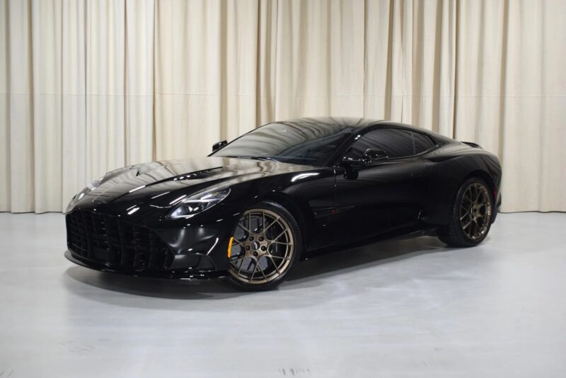 A stunning black Aston Martin Vanquish with bronze wheels is parked indoors in front of beige curtains.