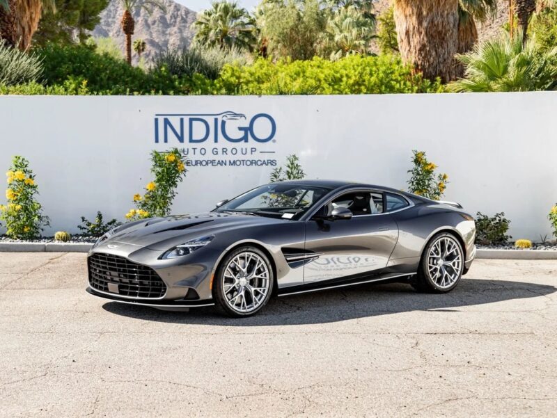 A dark gray Aston Martin Vanquish coupe is parked on pavement in front of a white wall with “INDIGO Auto Group” signage and green landscaping in the background, available now for sale.