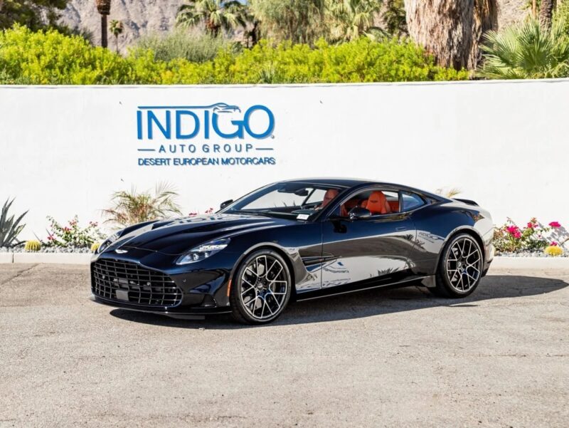 A black Aston Martin Vanquish sports car is parked in front of a white wall with the Indigo Auto Group logo and palm trees in the background, available now for sale.