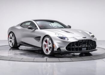 A silver Aston Martin Vanquish with a sleek design, low profile, and distinctive front grille is displayed in a studio with a white background, showcasing its powerful V12 engine.