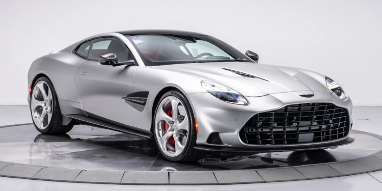 A silver Aston Martin Vanquish with a sleek design, low profile, and distinctive front grille is displayed in a studio with a white background, showcasing its powerful V12 engine.