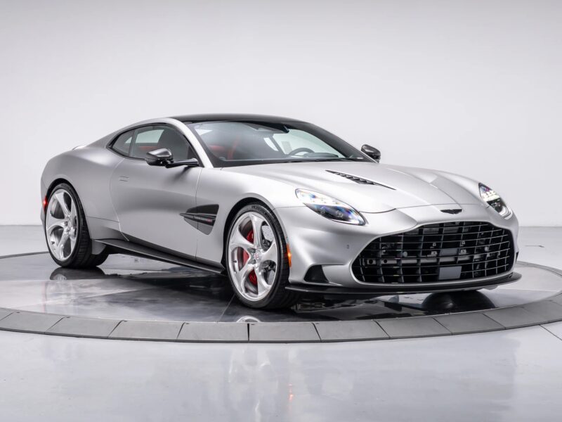 A silver Aston Martin Vanquish with a sleek design, low profile, and distinctive front grille is displayed in a studio with a white background, showcasing its powerful V12 engine.