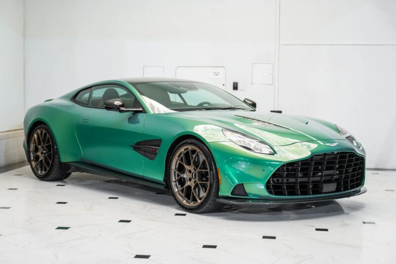 A green Aston Martin Vanquish two-door sports car with bronze alloy wheels is parked indoors on a white marble floor with black accents.