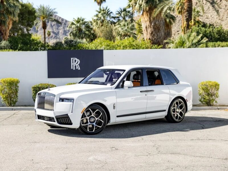 A white Rolls-Royce Cullinan, epitomizing pinnacle luxury, is parked in front of a white wall with palm trees and mountains as the backdrop. The SUV boasts a sleek design and prominent front grille, making it a true masterpiece of elegance.