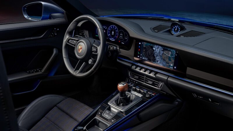 Interior view of a sports car featuring a steering wheel, digital dashboard, manual gear stick, and touchscreen display showing a map. The seats have plaid upholstery.