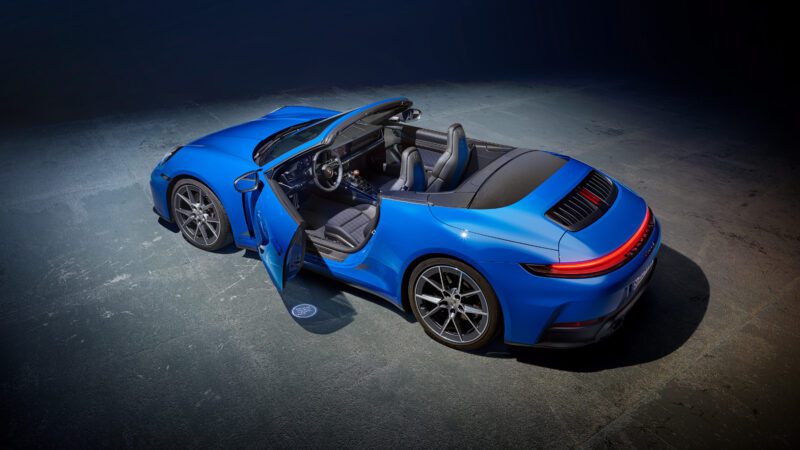 Blue convertible sports car with an open driver's door, viewed from above on a paved surface.