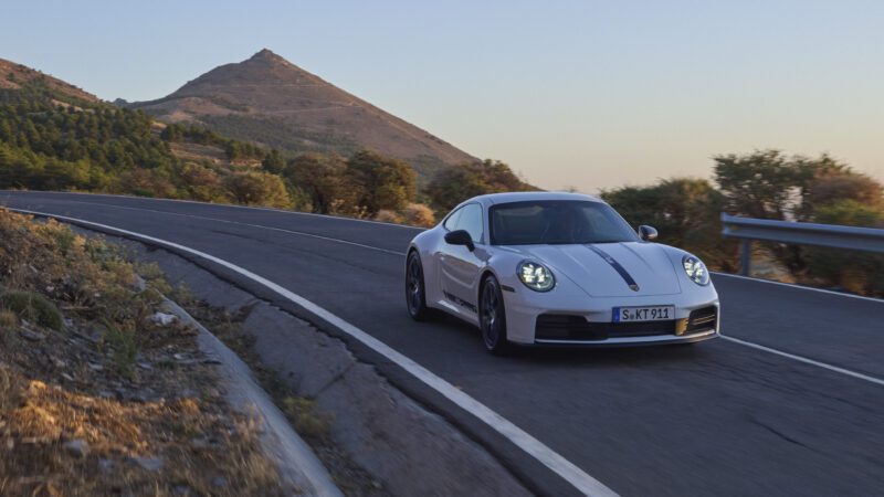 A white sports car driving on a winding mountain road with trees and a hill in the background.