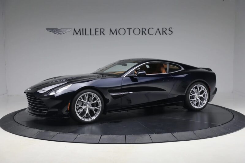 A dark blue Aston Martin Vanquish coupe with a powerful V12 engine is displayed in a showroom with "Miller Motorcars" on the wall behind it.