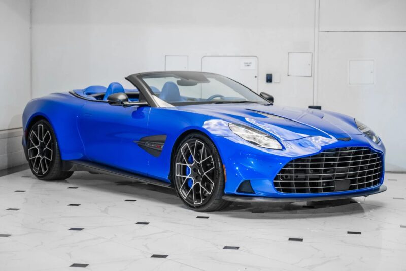An Aston Martin Vanquish V12 blue convertible sports car with black wheels is parked indoors on a white tile floor.
