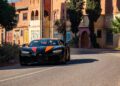 A sleek black hypercar with vibrant orange accents navigates a curved road, set against the backdrop of colorful traditional-style buildings and tall trees in Morocco.