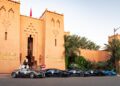 A row of luxury sports cars, including a sleek Bugatti hypercar, is parked in front of the Berbere Palace, an adobe-style building with palm trees nearby—a scene straight out of a Grand Tour.