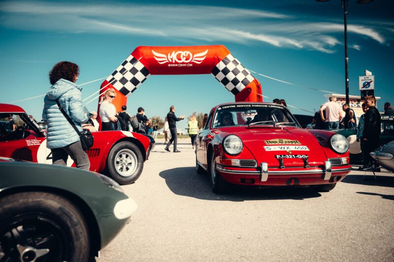 In 2024, classic cars and enthusiasts gathered at an outdoor event under a red and white arch with checkered flags, celebrating the Modena Cento Ore.