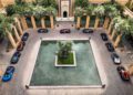 Aerial view of a courtyard in Morocco features a central pool enveloped by lush greenery and several parked luxury hypercars, including a stunning Bugatti.