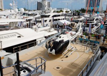 A luxury yacht with a helipad, showcasing a helicopter, is docked at a bustling marina teeming with other opulent vessels. As part of the prestigious 2024 Fort Lauderdale International Boat Show, onlookers are captivated by the display as they wander through this maritime spectacle.