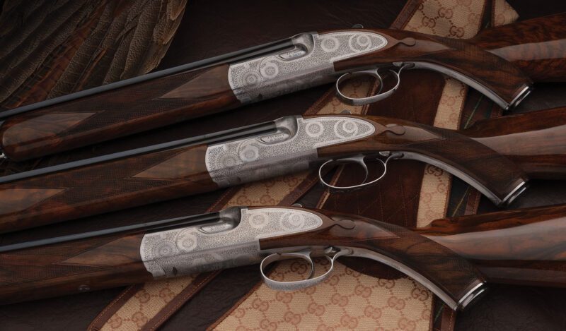 A close-up of three engraved double-barrel shotguns with wooden stocks, showcased on a leather surface, captures a sense of elegance akin to a finely curated Concours d'Elegance.