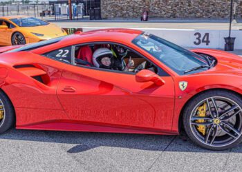 A 94-year-old sits inside a red Ferrari 488 GTB on the racetrack, clutching a phone. An orange sports car looms in the background, capturing the essence of a lifelong racing dream coming to life.