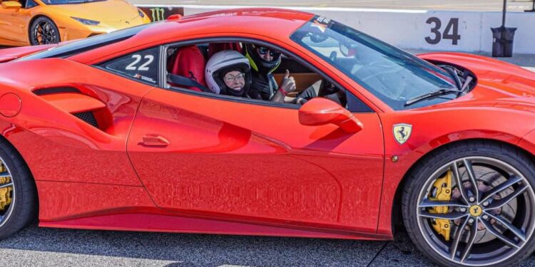 A 94-year-old sits inside a red Ferrari 488 GTB on the racetrack, clutching a phone. An orange sports car looms in the background, capturing the essence of a lifelong racing dream coming to life.